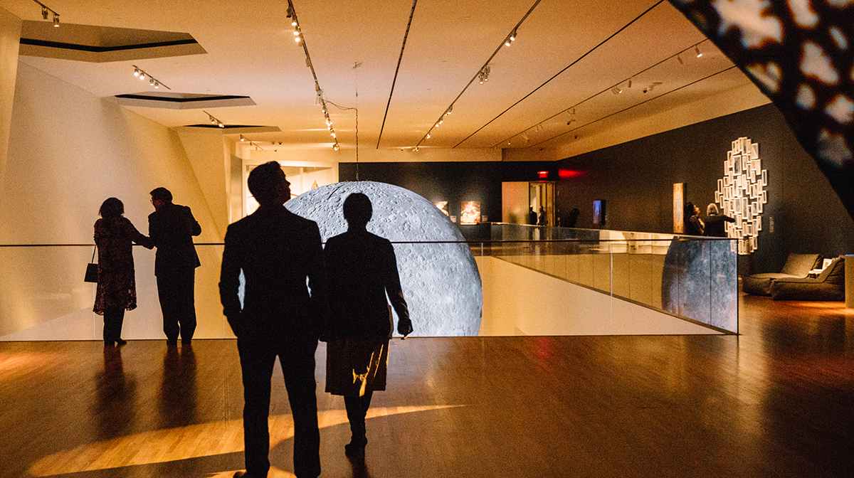Several pairs of people look at displays in the exhibition, including an illuminated sculpture of the moon.